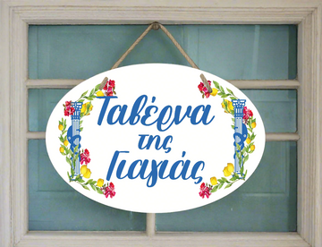 A decorative ceramic plaque with a white background, featuring a floral wreath and the text 'Taverna Tis Yiayias hanging by a wooden rope.