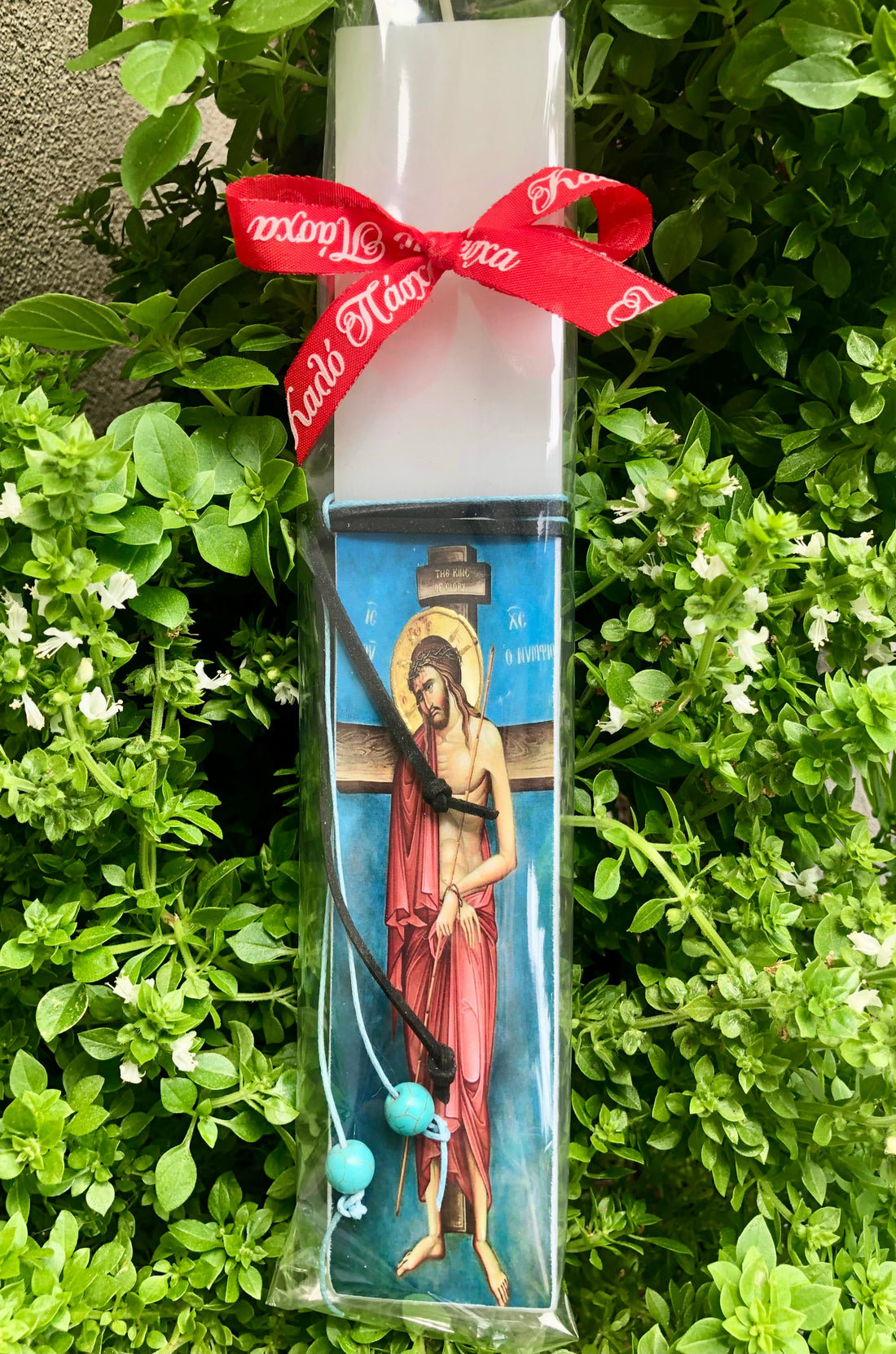 A packaged Easter candle with a depiction of Jesus Christ in red robes, adorned with a blue and gold design, wrapped in clear plastic with a red ribbon at the top.