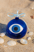 Blue glass ornament with an evil eye or mati design on a sandy surface with shells