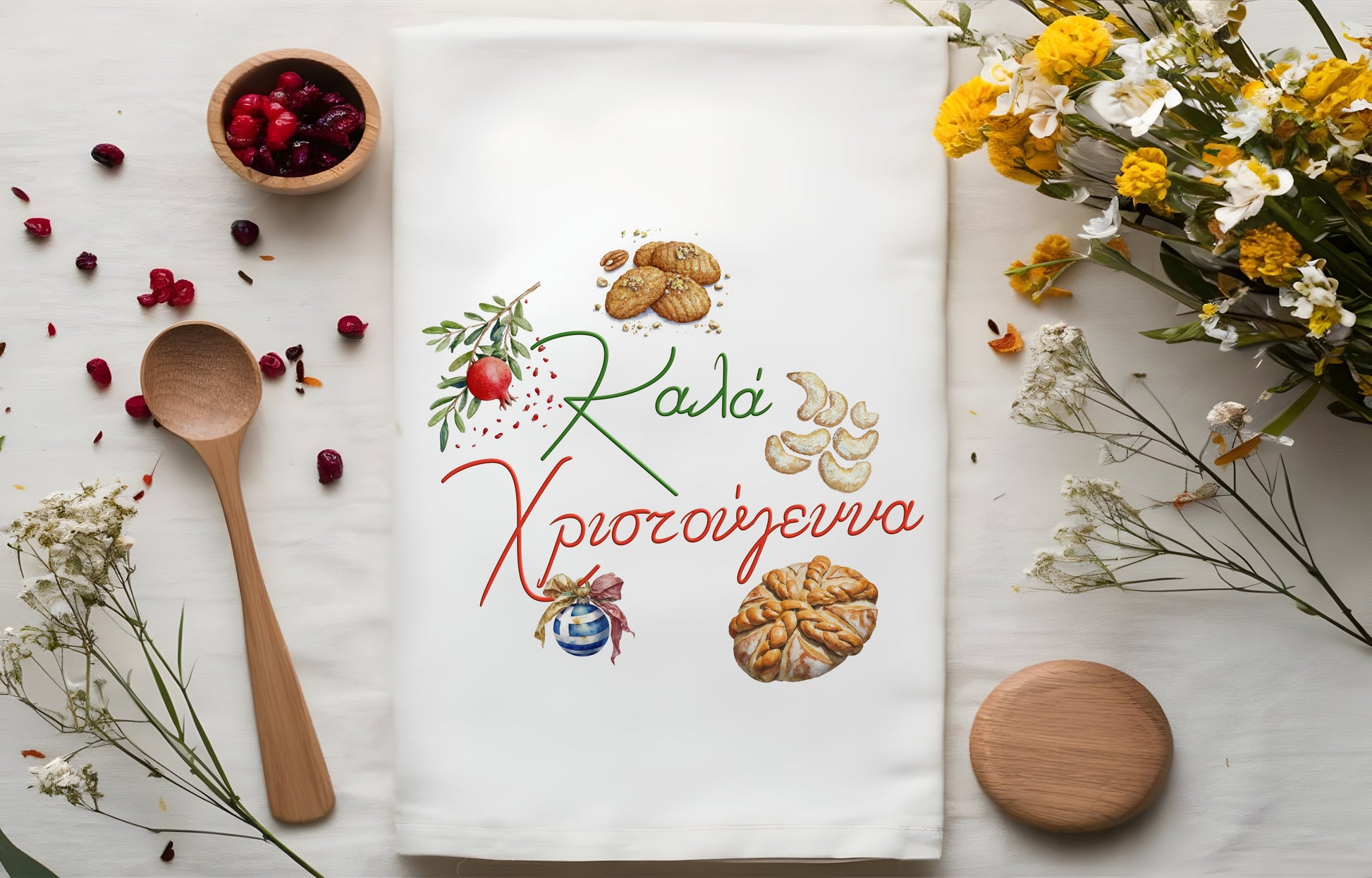 Decorative tea towel with greek sweets food-themed design and text, surrounded by wooden spoon, bowl of berries, and flowers.