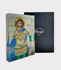 A hand-painted icon of Saint Artemios, depicted in a vibrant blue and gold robe, mounted on a black plexiglass cover with a decorative diamond-shaped logo on the front.