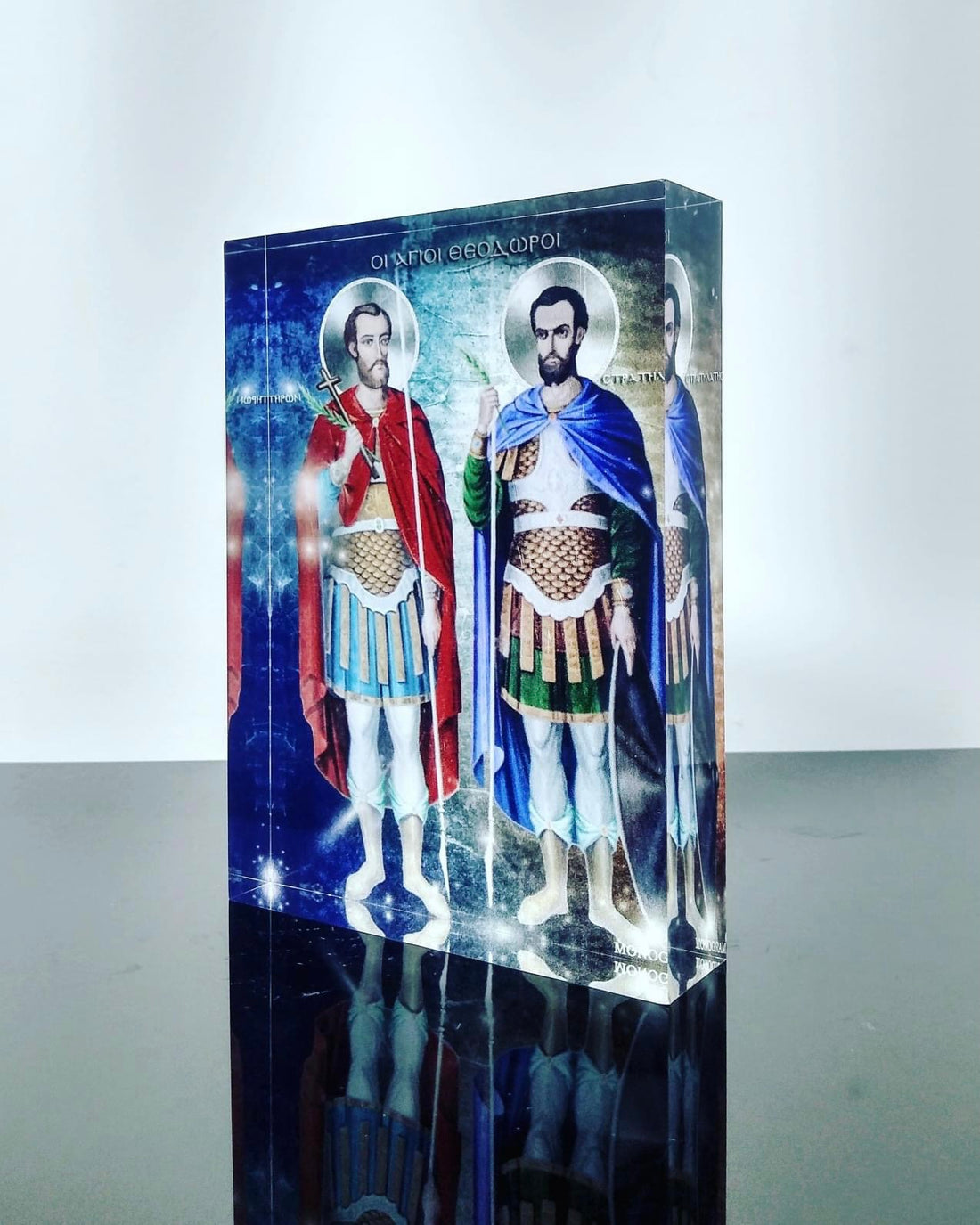 A plexiglass icon featuring two saints, Theodoros, with a blue and red robe and another in green and blue, standing side by side against a blue background.