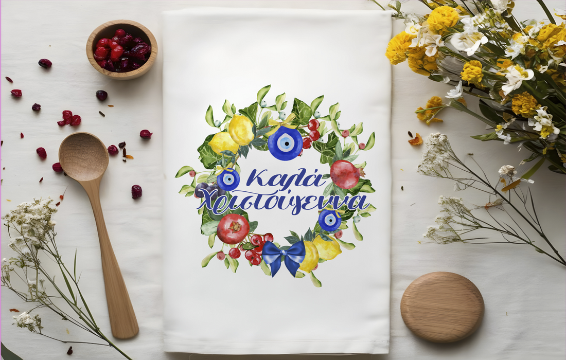 A folded white tea towel with a colorful Christmas themed design featuring Greek characters, pomegranates, and flowers, placed on a white surface with a wooden spoon and decorative flowers around it.