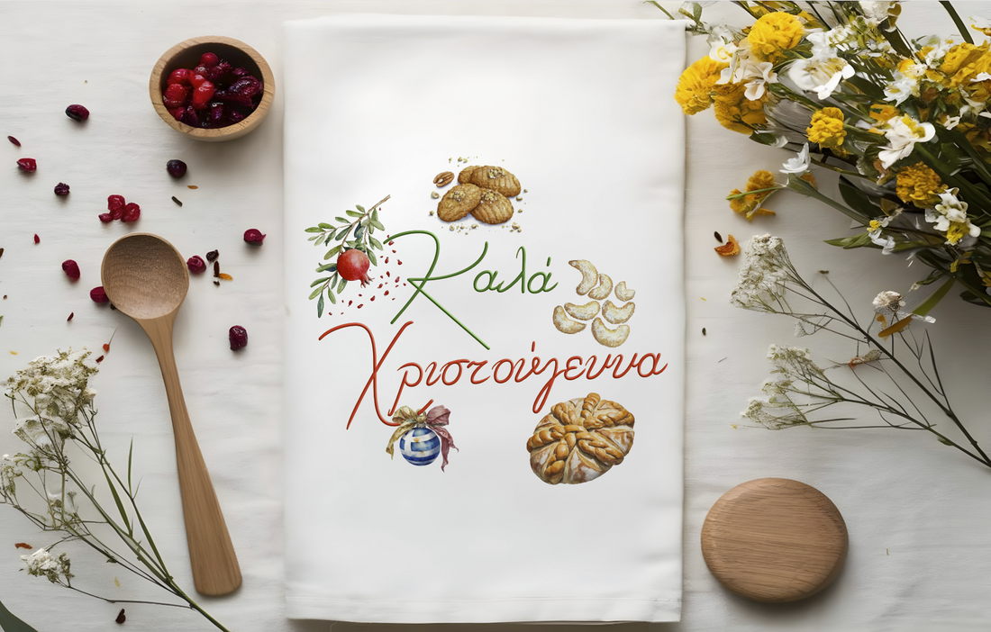 A folded white tea towel with a colorful Christmas themed design featuring Greek characters, pomegranates, and flowers, placed on a white surface with a wooden spoon and decorative flowers around it.