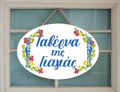 A decorative ceramic plaque with a white background, featuring a floral wreath and the text 'Taverna Tis Yiayias hanging by a wooden rope.