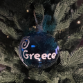 Blue glass Christmas bauble with white Greece lettering and an Ionic column design, hanging on a green Christmas tree by a jute loop.