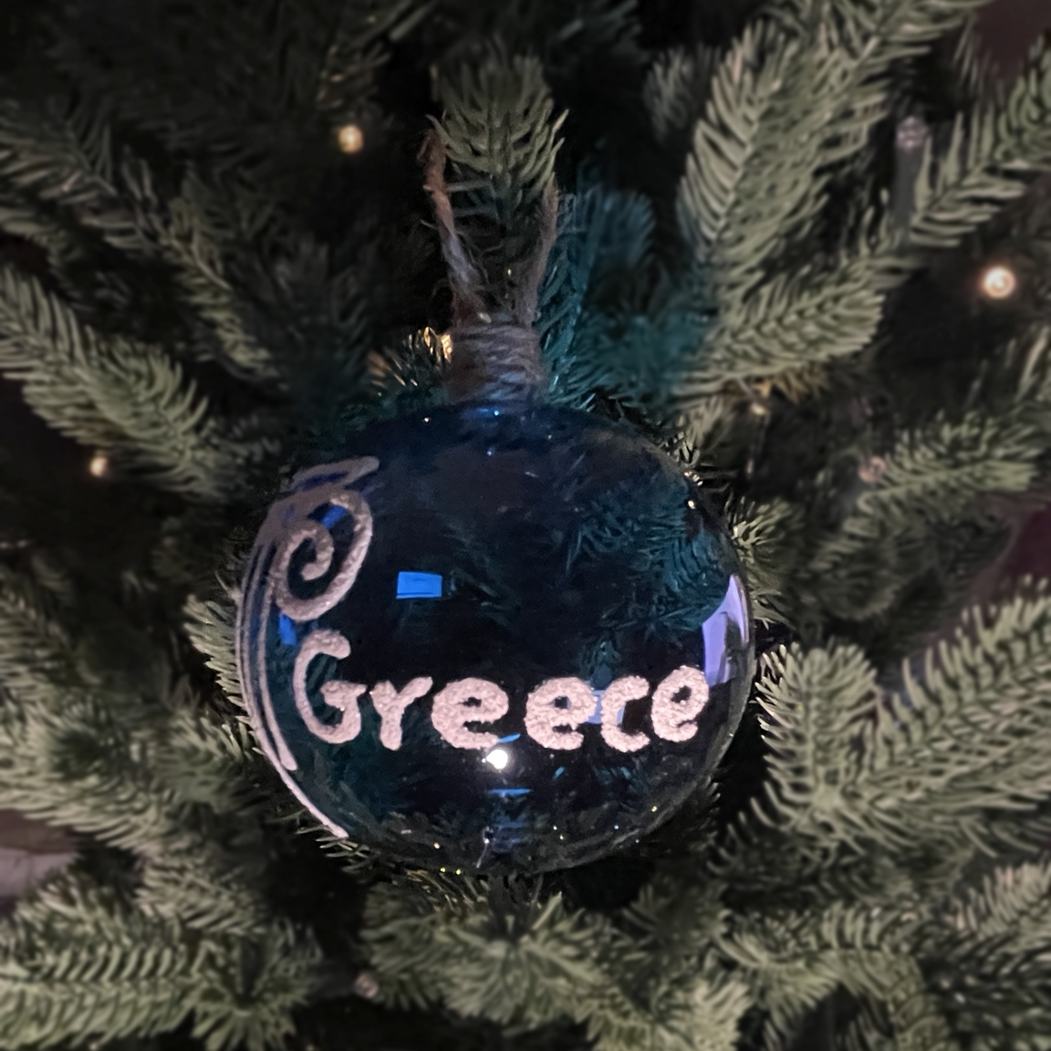 Blue glass Christmas bauble with white Greece lettering and an Ionic column design, hanging on a green Christmas tree by a jute loop.