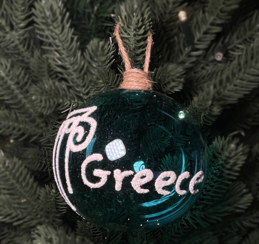 Turquoise glass Christmas bauble with white Greece lettering, an Ionic column design and a jute hanging loop, nestled among green Christmas tree branches.