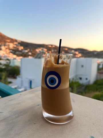 Frappe Coffee with a straw on a ledge overlooking a scenic town at sunset.
