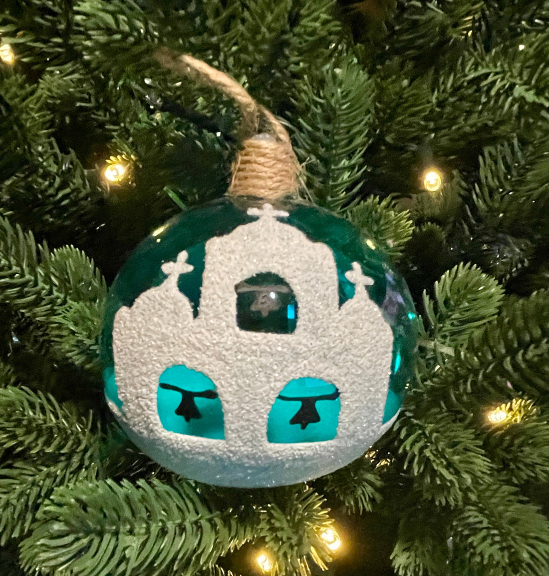 Hand-blown glass Christmas bauble with turquoise and white Greek churches hanging on green Christmas tree branches.