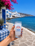 Hand holding a traditional Greek ouzo glass by the sea with a whitewashed coastal village in the background.