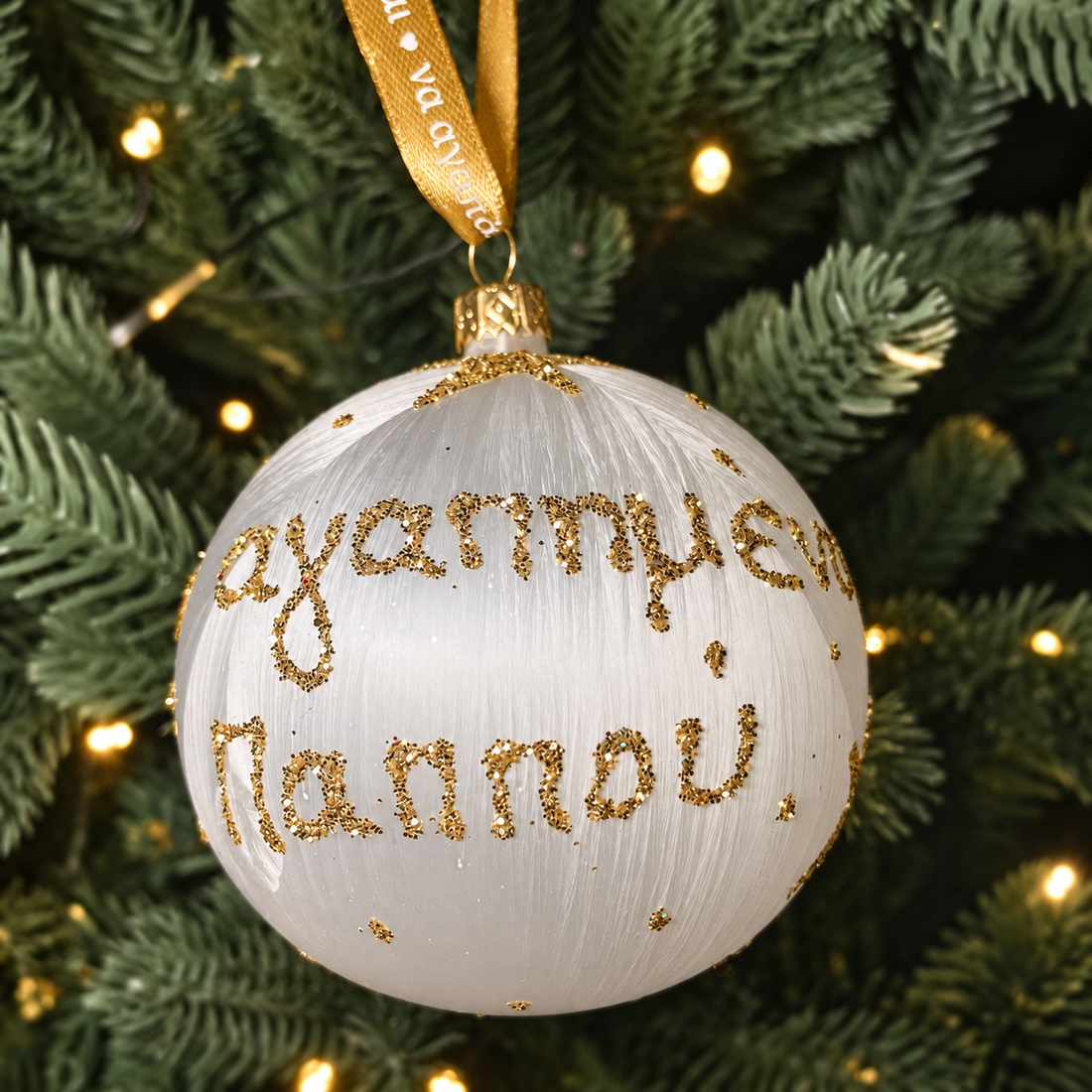 White Greek Yiayia Christmas bauble with gold glitter Greek lettering, star details and gold ribbon hanging on a Christmas tree.