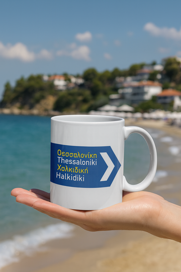White mug with blue text and arrow design held by a hand on a beach.  Thessaloniki and Halkidiki