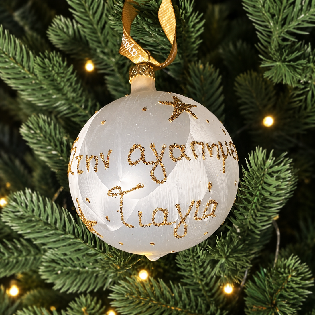 White Greek Yiayia Christmas bauble with gold glitter Greek lettering, star details and gold ribbon hanging on a Christmas tree.