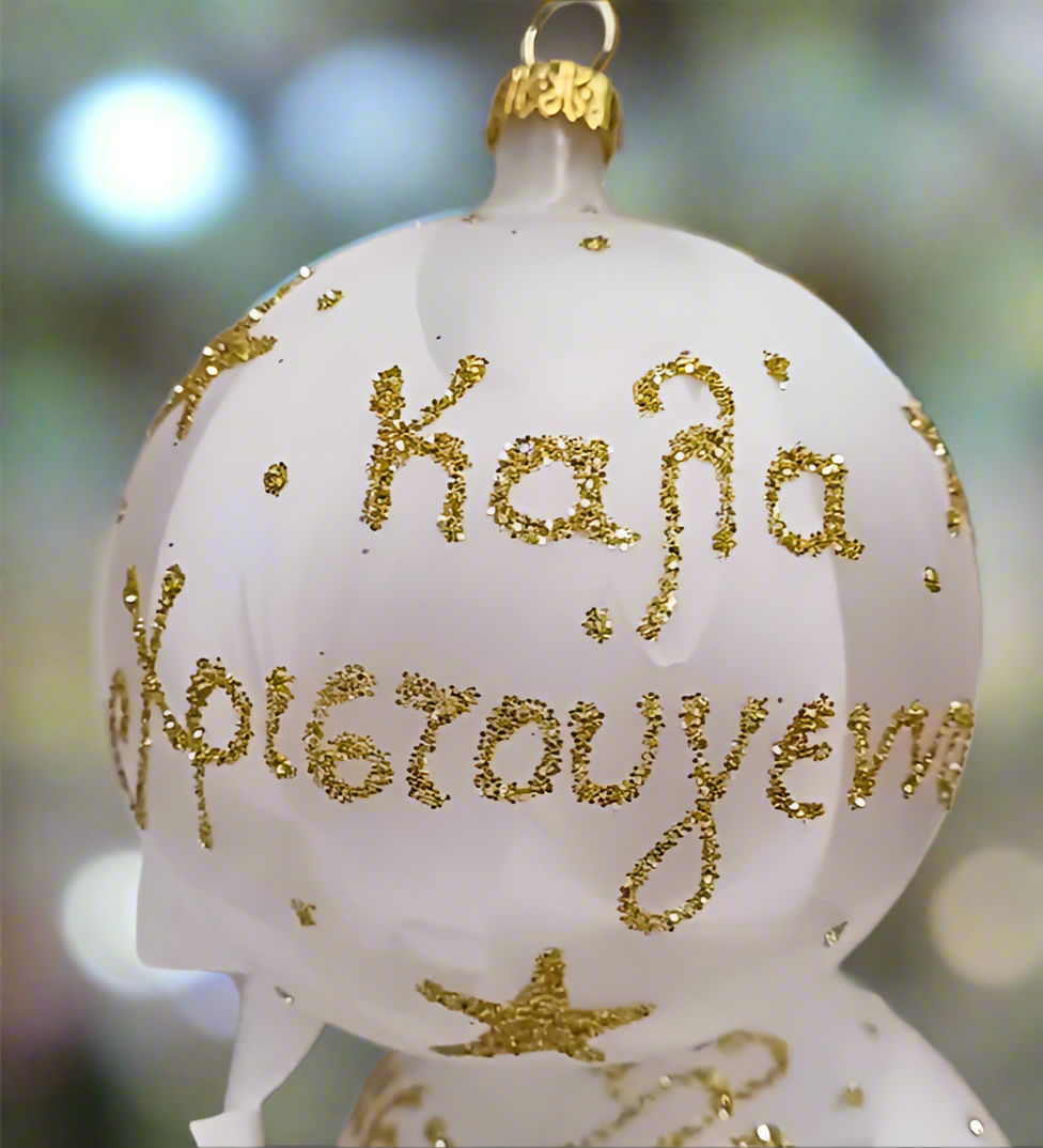 White glass Christmas bauble with gold glitter Καλά Χριστούγεννα lettering and stars, hanging from a gold ribbon on a green Christmas tree.