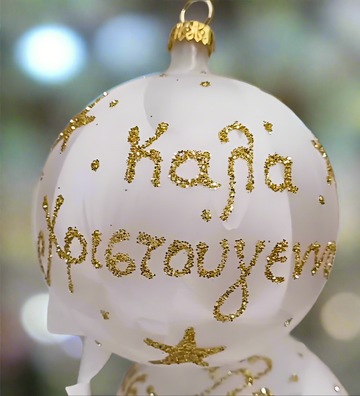 White glass Christmas bauble with gold glitter Καλά Χριστούγεννα lettering and stars, hanging from a gold ribbon on a green Christmas tree.