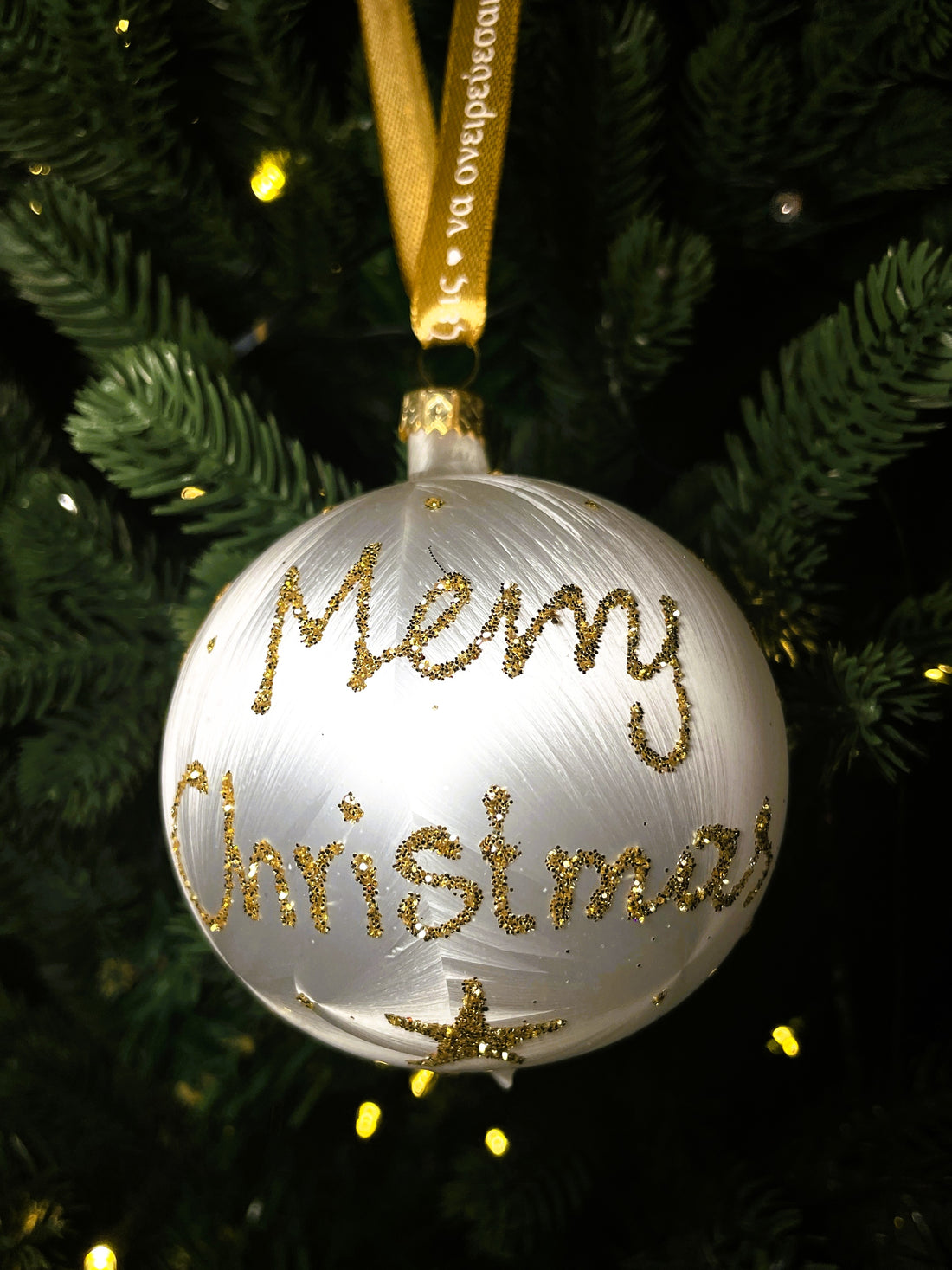 White glass bauble with gold glitter Merry Christmas lettering and stars, hanging from a gold ribbon on a green Christmas tree.