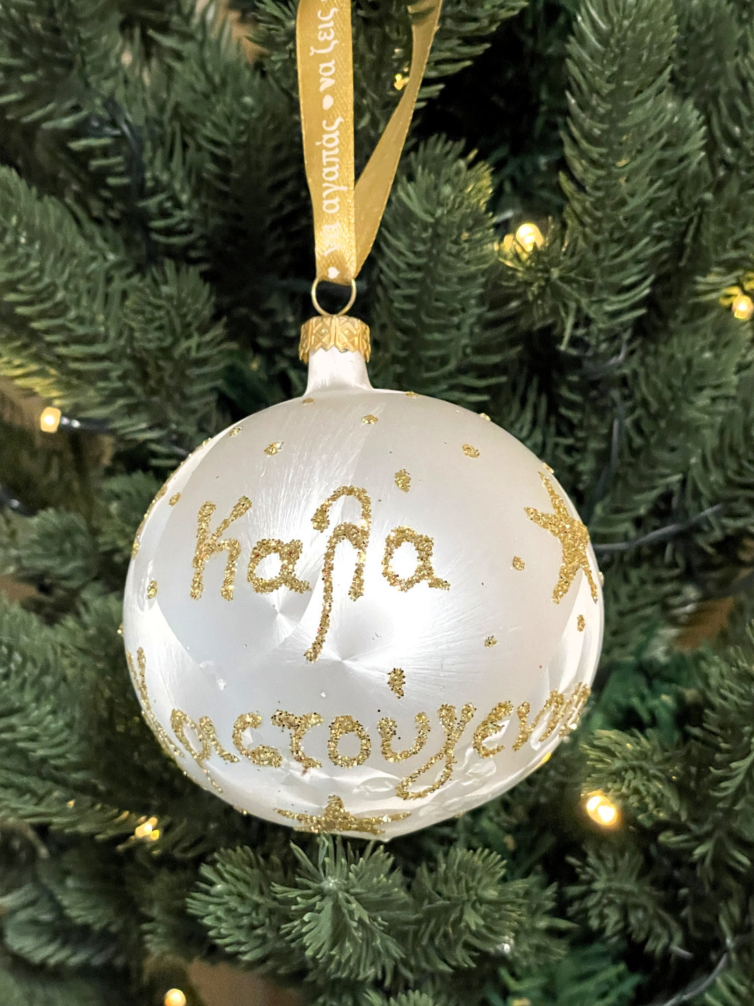 White glass bauble with gold glitter Merry Christmas lettering and stars, hanging from a gold ribbon on a green Christmas tree.