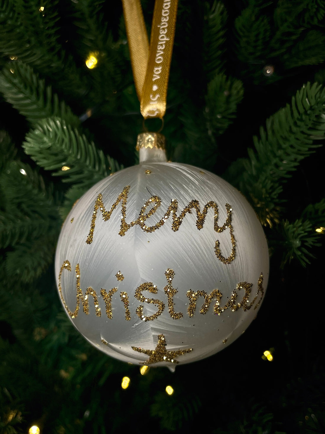 White glass Christmas bauble with gold glitter Καλά Χριστούγεννα lettering and stars, hanging from a gold ribbon on a green Christmas tree.