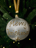White glass bauble with gold glitter Merry Christmas lettering and stars, hanging from a gold ribbon on a green Christmas tree.