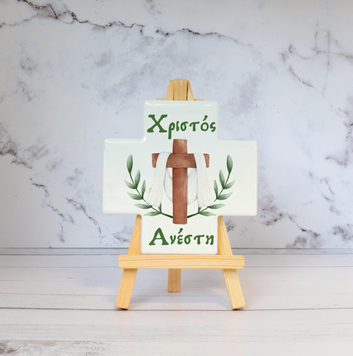 A ceramic cross with a stand, featuring a glossy finish with green leaf patterns and text, displayed on a wooden easel against a marble background.