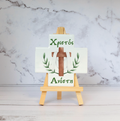 A ceramic cross with a stand, featuring a glossy finish with green leaf patterns and text, displayed on a wooden easel against a marble background.