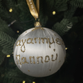 White glass Pappou Christmas bauble with gold glitter Greek lettering and a gold ribbon, hanging on a green Christmas tree.