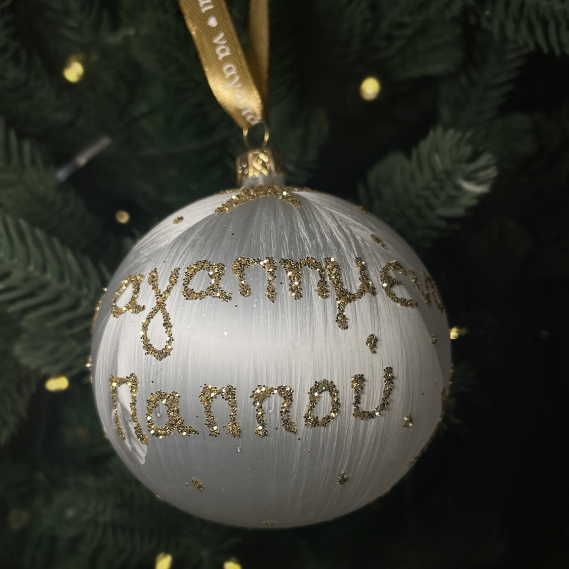 White glass Pappou Christmas bauble with gold glitter Greek lettering and a gold ribbon, hanging on a green Christmas tree.