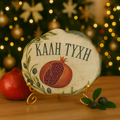 Greek pomegranate good luck plate with “ΚΑΛΗ ΤΥΧΗ” on a gold stand, displayed in front of a Christmas tree with warm festive lights.