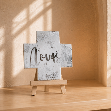 White ceramic Greek Orthodox name cross with silver detailing displayed on a wooden stand in a warm neutral prayer corner setting