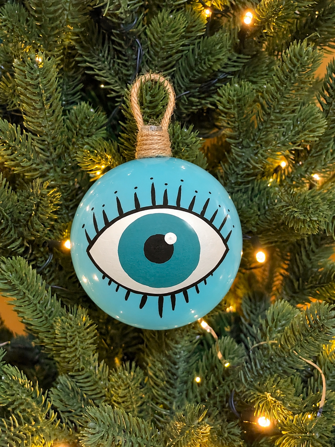 Sky blue Greek evil eye Christmas ornament with hand-painted eye and black eyelashes, displayed among Christmas tree lights.