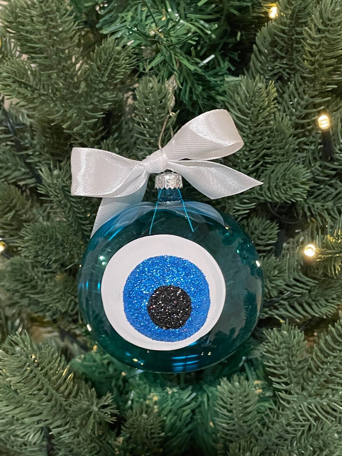 Turquoise glass Greek evil eye Christmas bauble with blue glitter mati design and white bow hanging on a Christmas tree.
