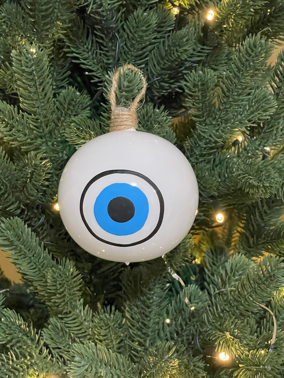 Turquoise glass Greek evil eye Christmas bauble with blue glitter mati design and white bow hanging on a Christmas tree.