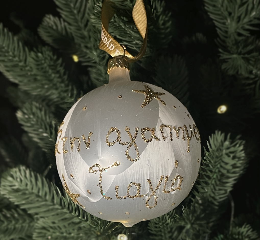 White glass Pappou Christmas bauble with gold glitter Greek lettering and a gold ribbon, hanging on a green Christmas tree.