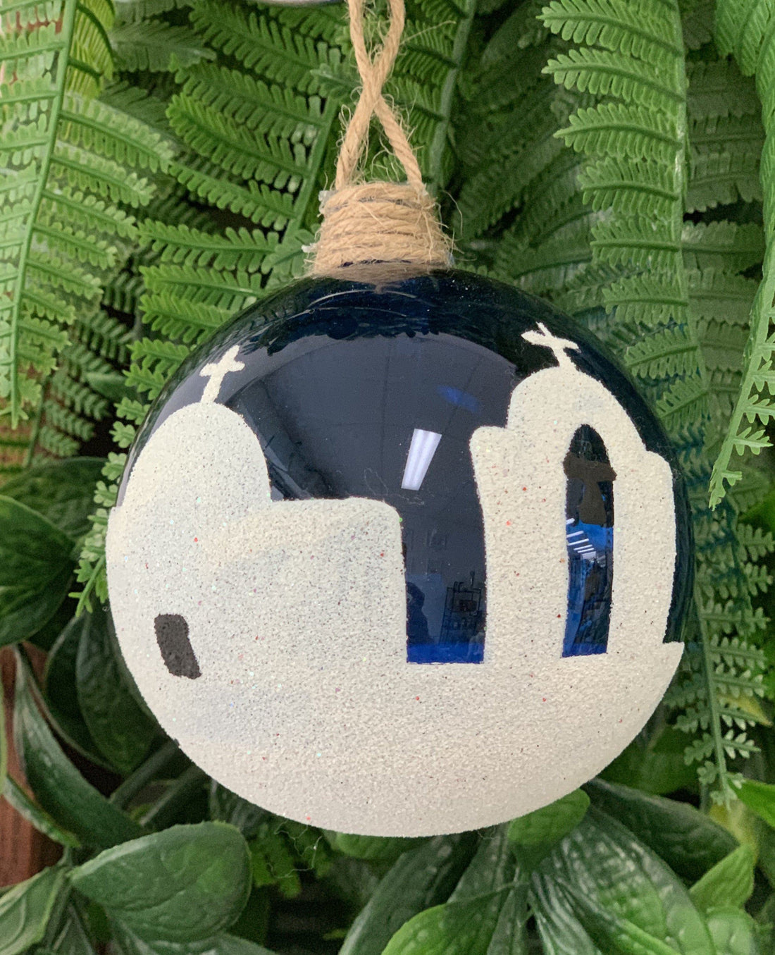 A hand blown glass bauble with a blue and white design, featuring a silhouette of a church, displayed against a green fern backdrop.