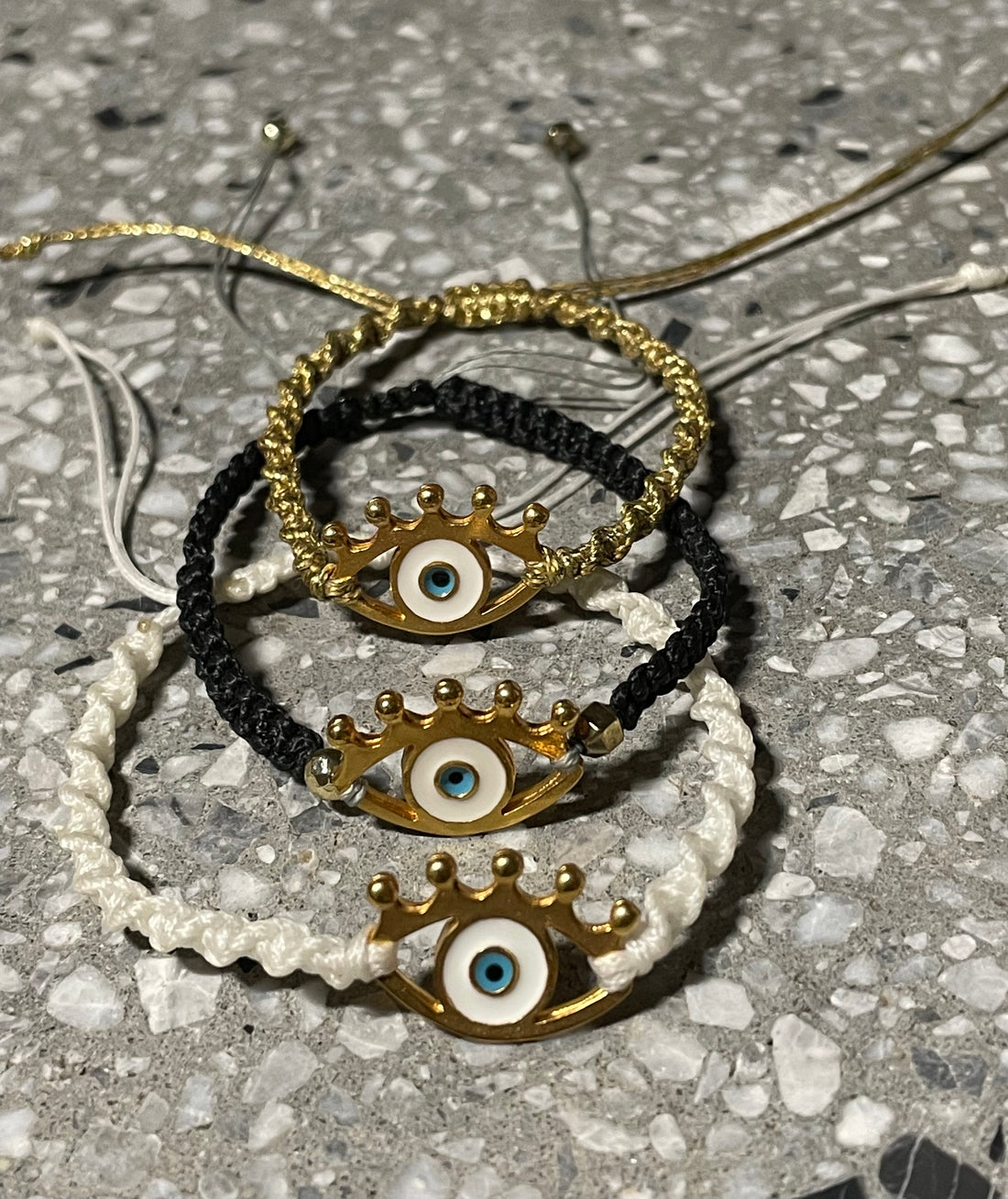 A set of macrame bracelets with evil eye pendants in white and gold colors, displayed on a textured background.