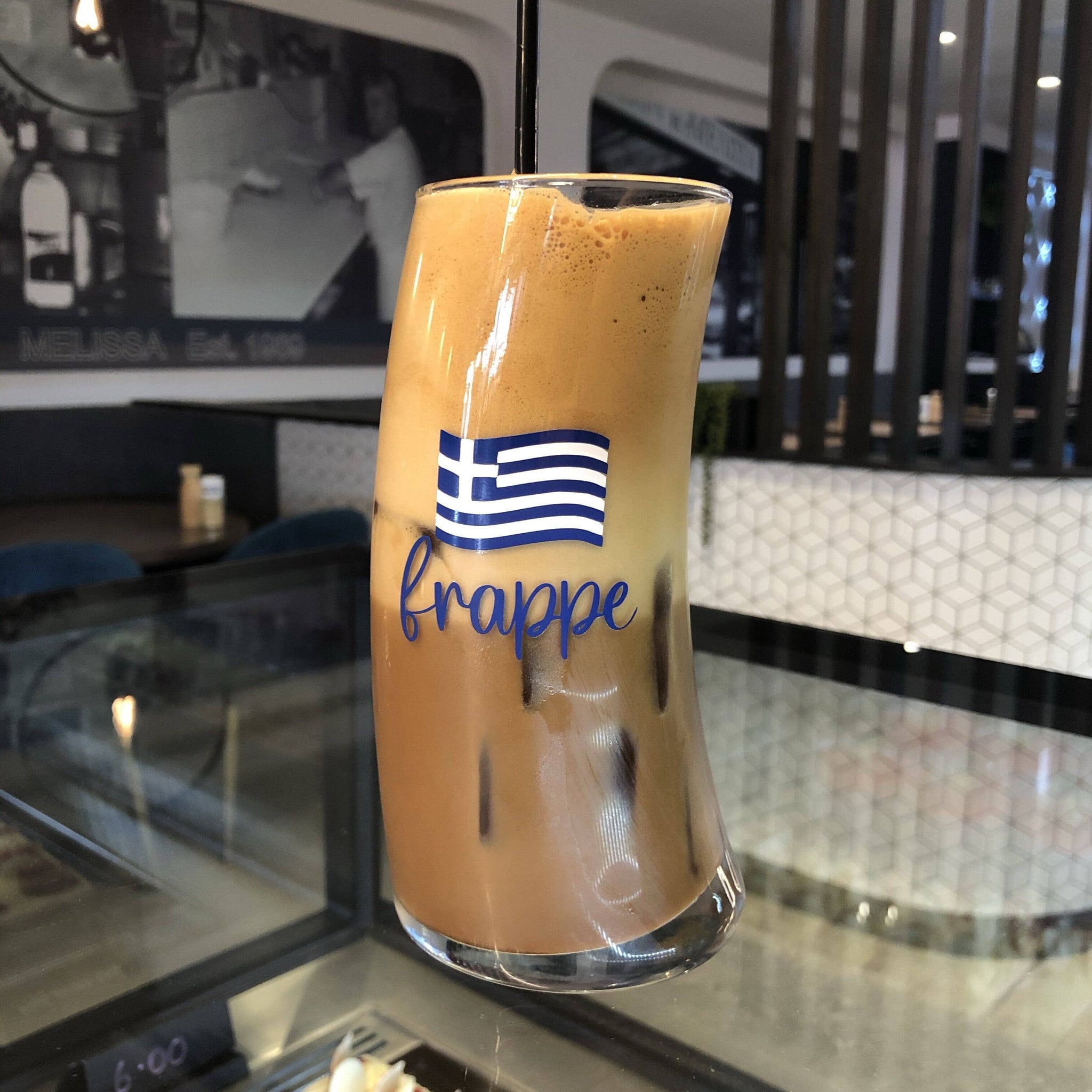 A glass of frappe with a Greek flag-themed design, served on a table in a cafe setting.
